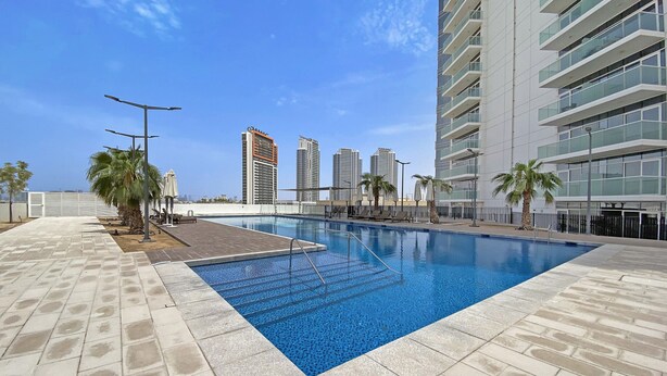 A swimming pool with a tiled deck, palm trees, and modern buildings in the background. - Chic Hotel-Style Studio in Damac Hills (Dubai)