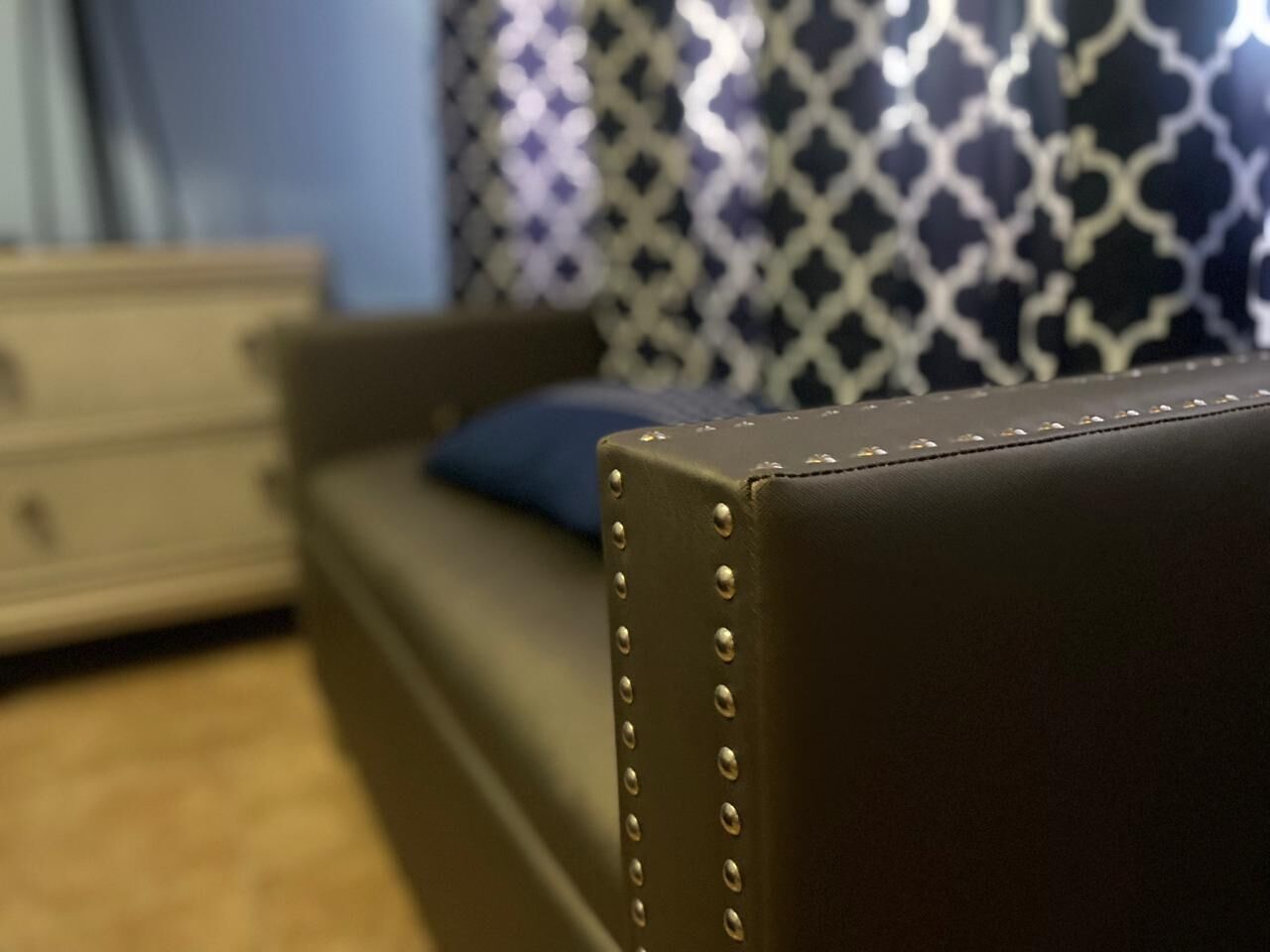 A close-up of a brown leather sofa with riveted edges, a blue pillow, and a patterned curtain in the background.