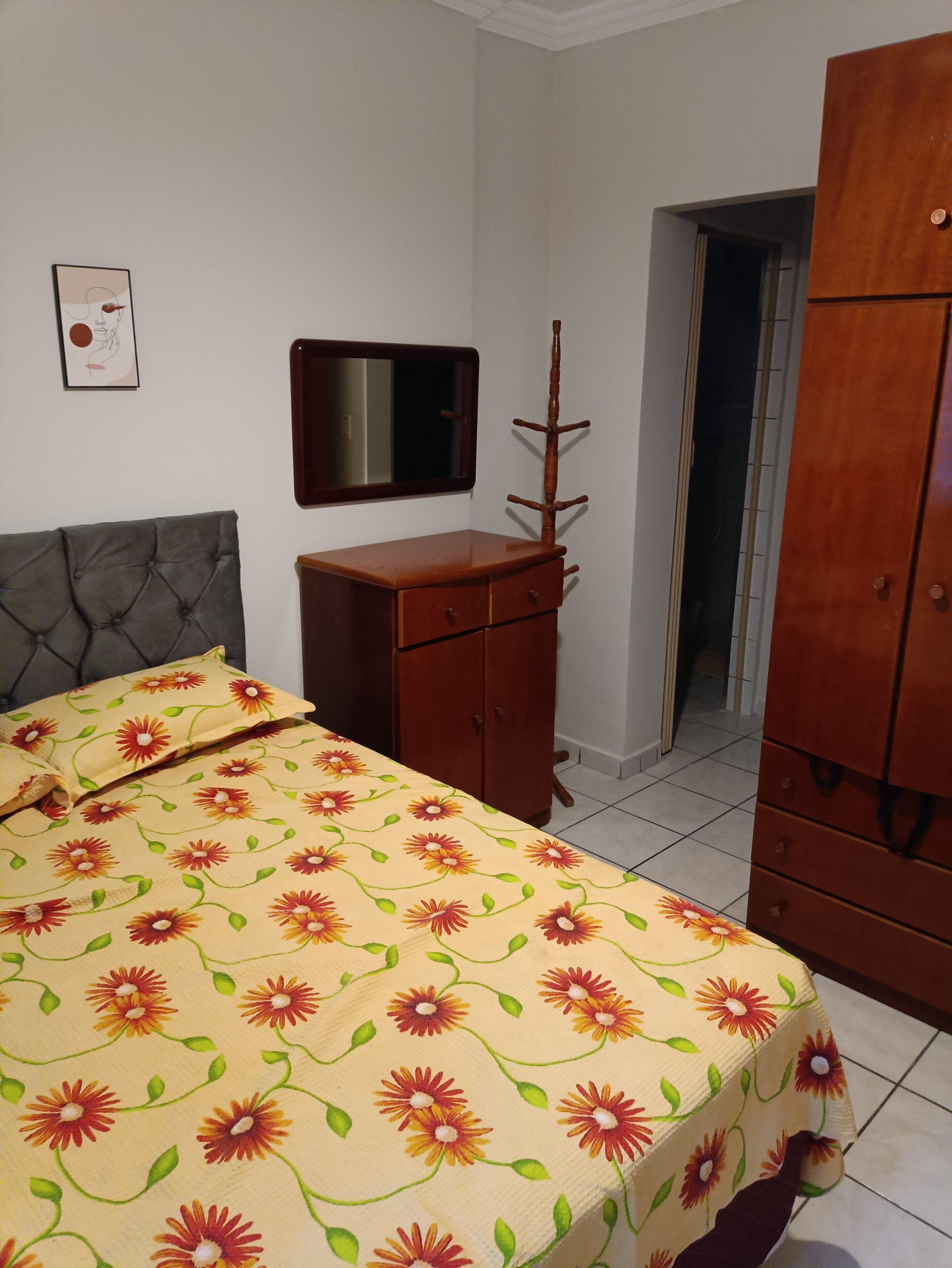 Um quarto com uma cama, cômoda de madeira, espelho e porta que dá para outro espaço.