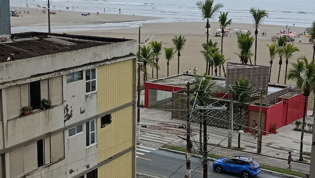 A multi-story building with balconies, a street with cars, and a beach in the background. - Ótimo Apartamento Vista Mar Pé na Areia (Praia Grande)