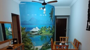A living room with a brown sofa, a dining table with chairs, a ceiling fan, and a mural of a tropical beach scene. - Ótimo Apartamento Vista Mar Pé na Areia (Praia Grande)
