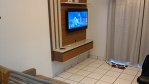A living room with a flat-screen TV mounted on a wooden wall, a sofa, a chair, and a small table. - Ótimo Apartamento Vista Mar Pé na Areia (Praia Grande)