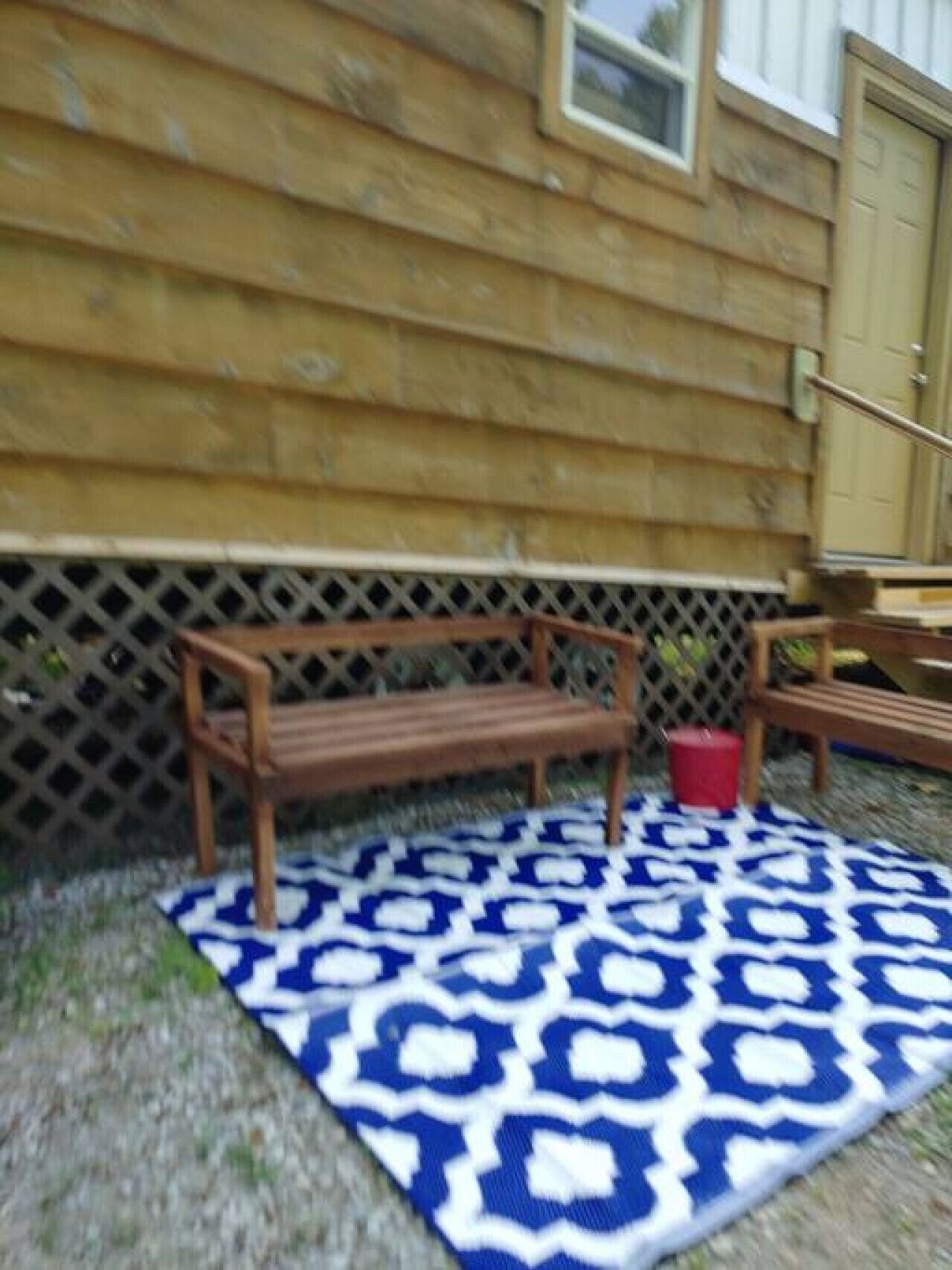 Un banc en bois et un seau rouge sont posés sur un tapis à motifs devant un bâtiment en bois, délimité par une clôture à treillis.