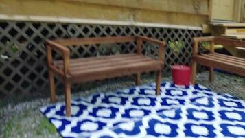 Un banc en bois et un seau rouge sont posés sur un tapis à motifs devant un bâtiment en bois, délimité par une clôture à treillis.