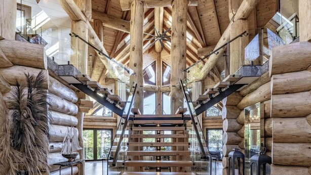 A log cabin interior with a central staircase, wooden beams, and large windows. - 7 bed 6 bath cottage on Lake Muskoka (Gravenhurst)