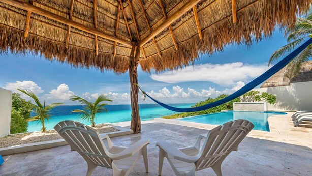 A poolside area with two white plastic chairs, a thatched roof, a hammock, and a view of the ocean. - Luxurious 5 BR Ocean Front Home. Epic Views! (Isla Mujeres)