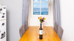 Un tavolo da pranzo con una bottiglia di vino e bicchieri, un vaso con fiori, e vista sul mare attraverso una finestra con tende.