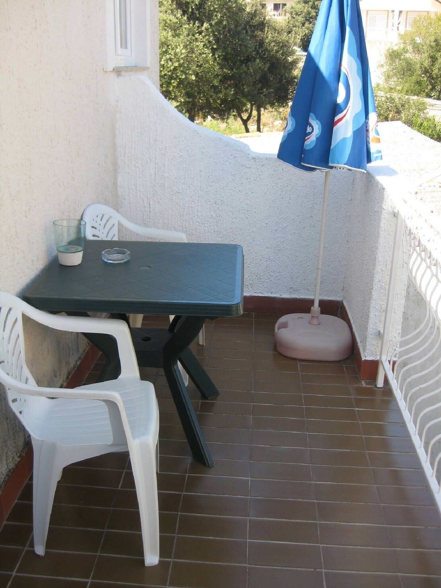 Pequeno pátio externo com mesa e duas cadeiras de plástico branco, um guarda-sol azul e um balde rosa para limpeza.