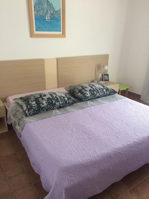 A neatly made bed with a purple bedspread and patterned pillows, a wooden headboard, a small bedside table with a lamp, and a framed picture of a sailboat on the wall.