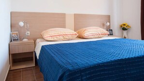 A neatly made bed with a blue bedspread, two striped pillows, a nightstand with a lamp, a framed picture, and a vase with sunflowers. - Apartments Paula - One Bedroom Apartment with Terrace 2 (Jezera)