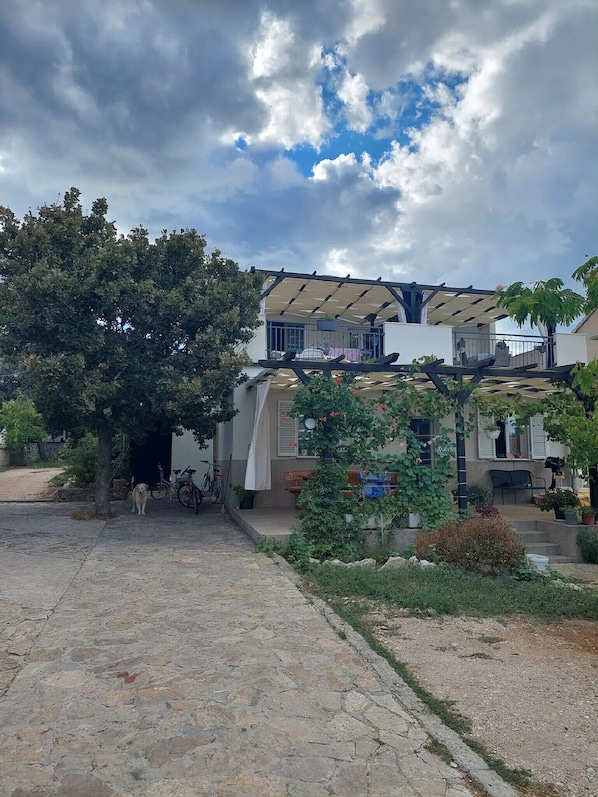 A two-story house with a balcony, surrounded by greenery and a paved driveway.