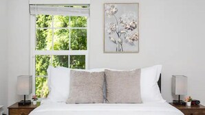 Une chambre avec une grande fenêtre, un lit avec une literie blanche et grise, une œuvre d’art florale encadrée au mur et des lampes de chevet.