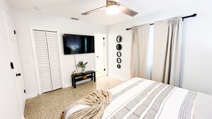 Een slaapkamer met een bed, een televisie, een plafondventilator, een kleine tafel met een plant en een deur met verstelbare luiken.