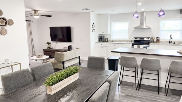 Een moderne keuken met witte kastjes, een centraal eiland met barkrukken, een eettafel met een plant als middelpunt en een flatscreen televisie aan de muur.