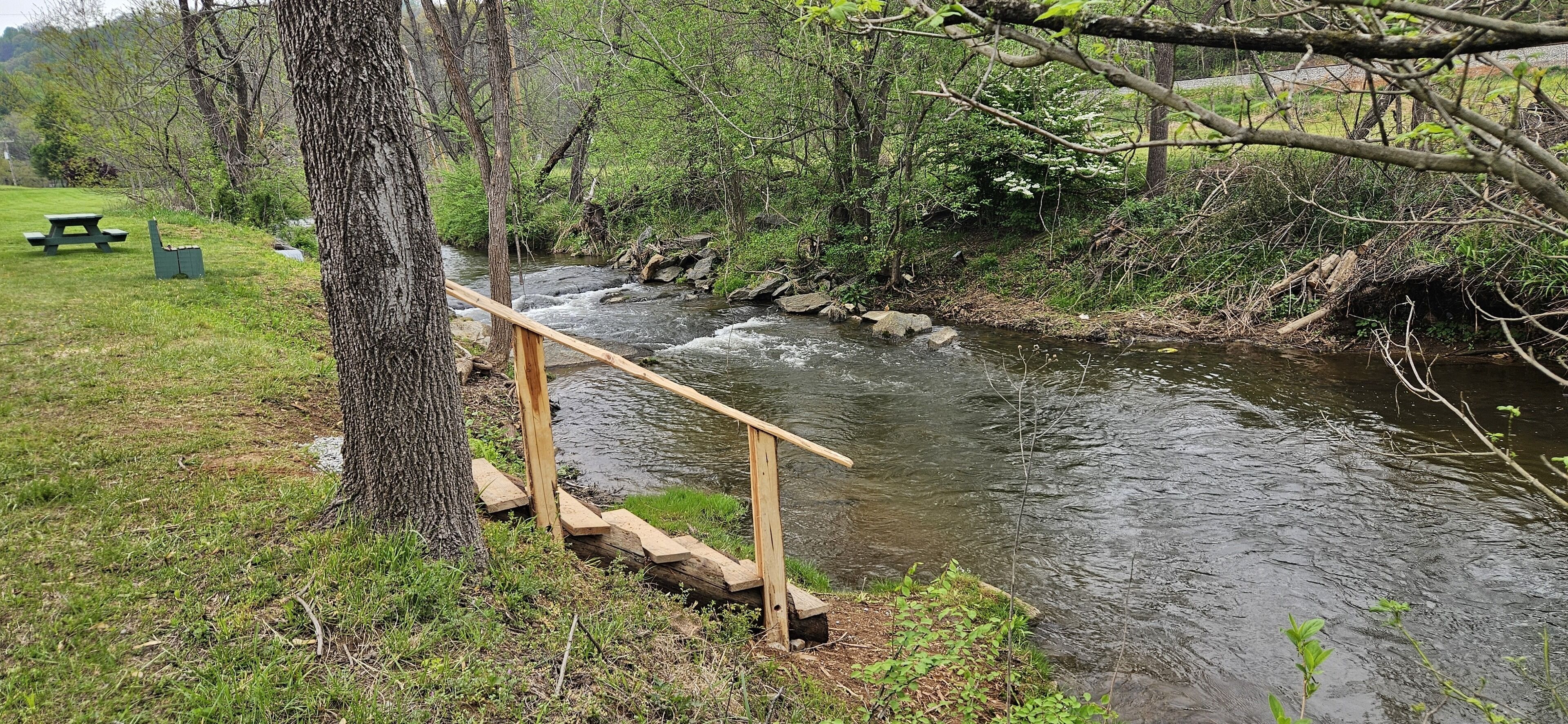 A wooden bridge over a river with a picnic table and benches nearby.