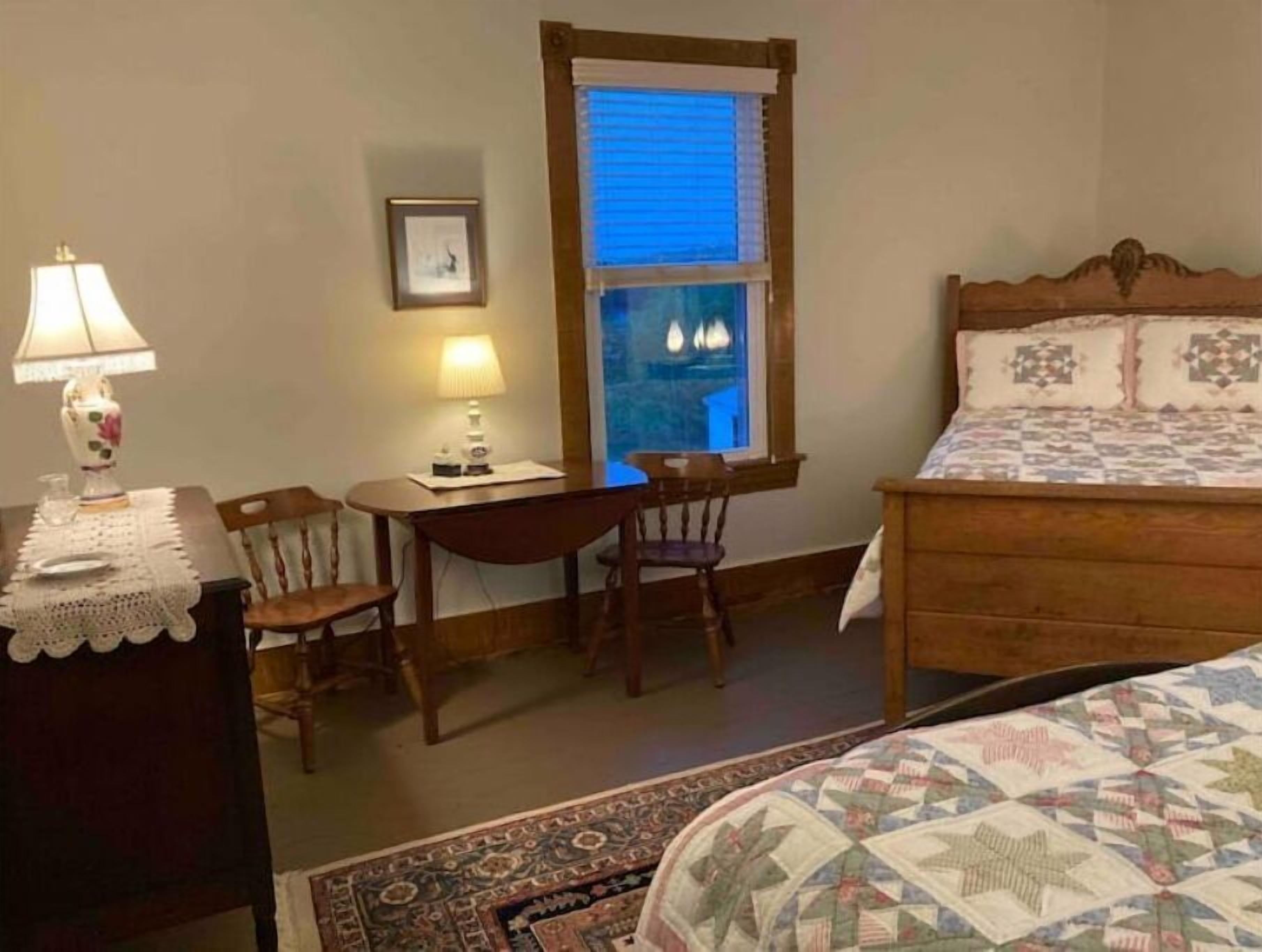 A bedroom with a bed, a desk with a chair, a lamp, a window with blinds, and a framed picture on the wall.