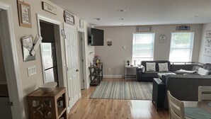 A spacious living area with a wooden floor, a sofa, a dining table, and a kitchen area with white cabinets and a refrigerator. - Cozy Country Cottage #2 (Miller Place)