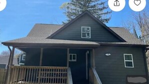 A two-story house with a dark exterior, a front porch with a wooden railing, and a small window on the second floor. - Cozy Country Cottage #2 (Miller Place)