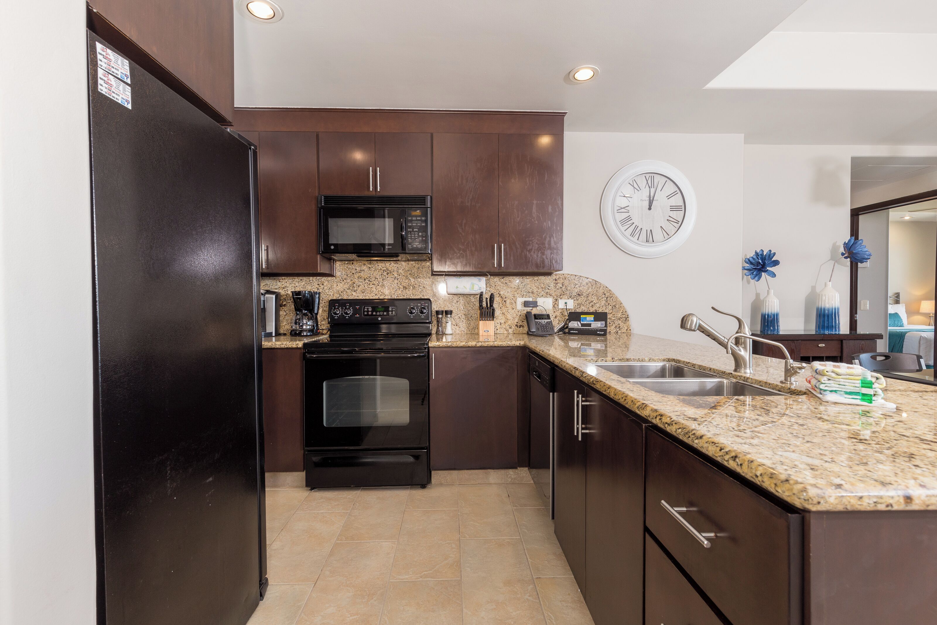 A modern kitchen with dark wood cabinets, a granite countertop, stainless steel appliances, and a wall clock.