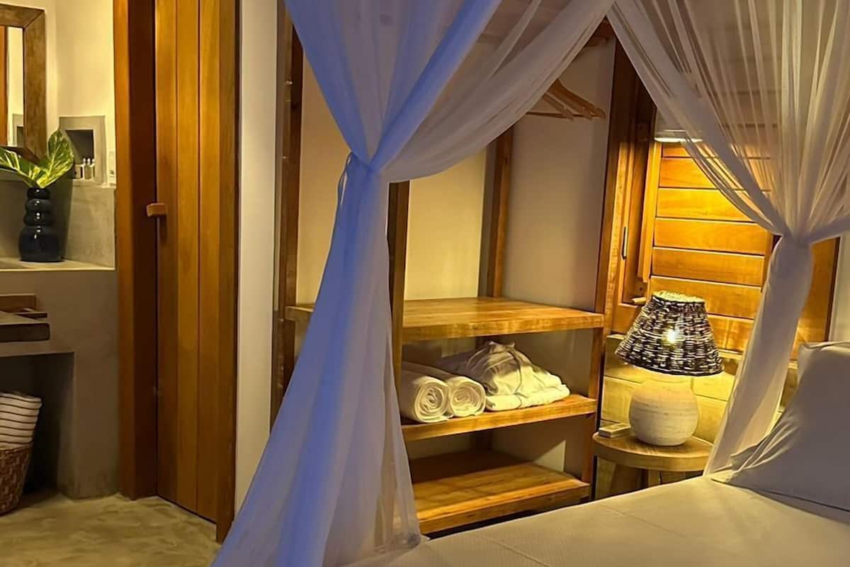 Een slaapkamer met een hemelbed, houten planken met opgevouwen handdoeken, een bedlampje en een potplant.