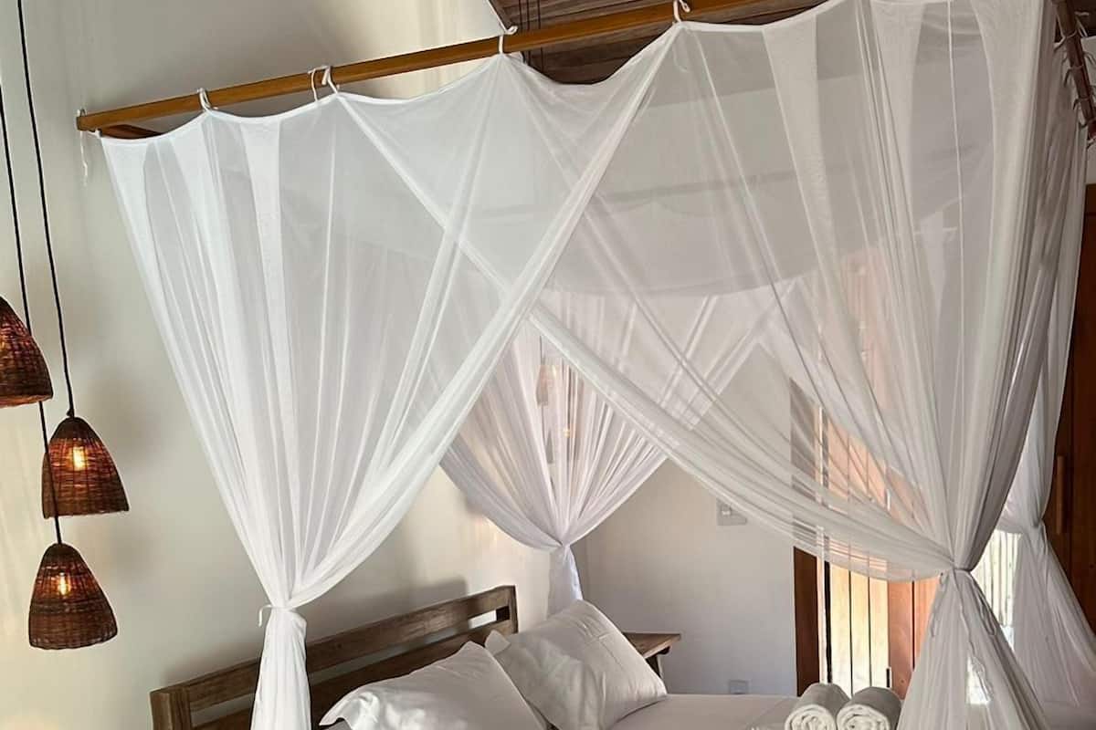 Een slaapkamer met een bed onder een witte hemel, een houten bankje en een geweven mat op de vloer.