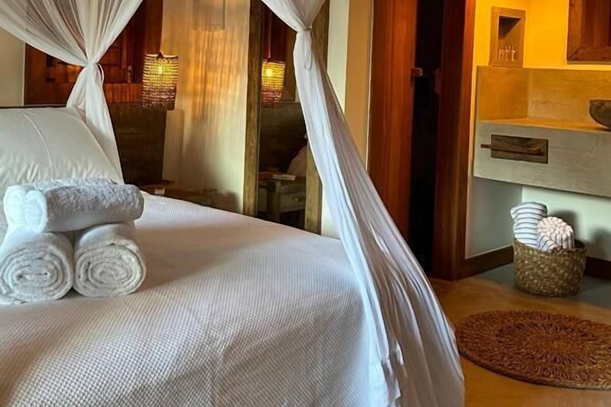 Een net opgemaakt bed met witte lakens en een hemelbed, een houten deur, een wandlamp en een geweven mand op de vloer.
