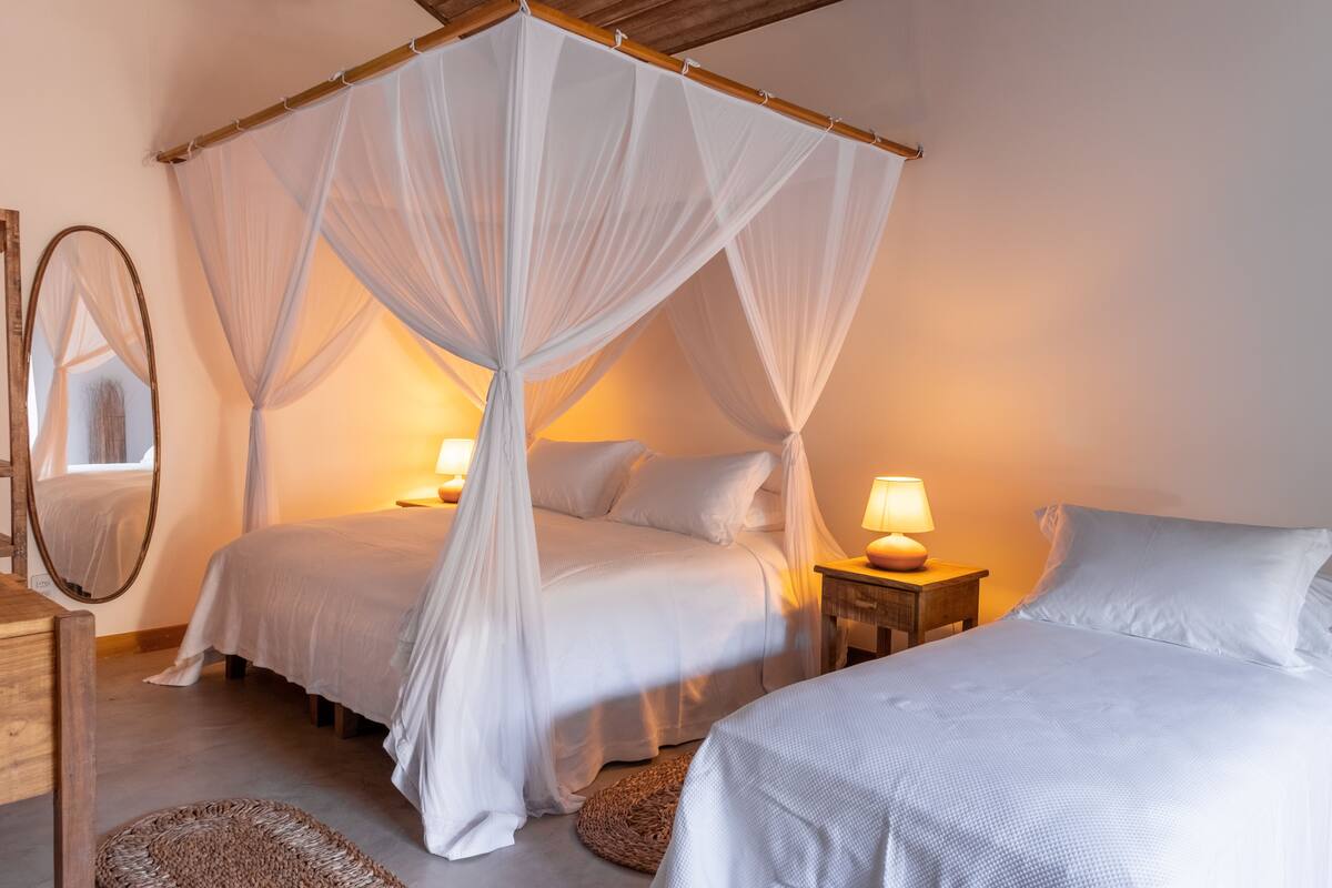 Een slaapkamer met een hemelbed, twee nachtkastjes met lampen, een rond geweven vloerkleed en een spiegel aan de muur.