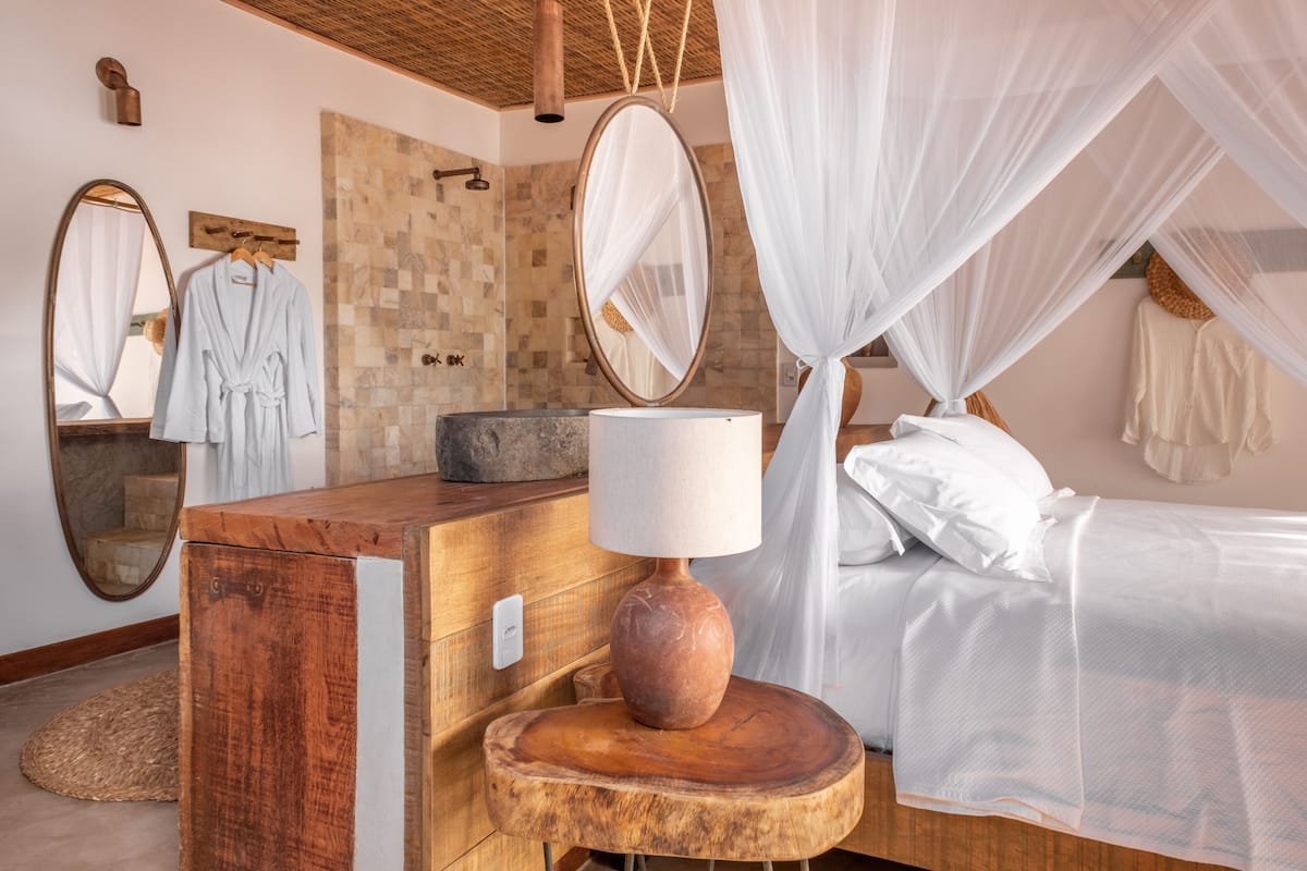 Een slaapkamer met een houten dressoir, een nachtkastje met een lamp, een bed met witte beddengoed, een hemelbed en een spiegel.