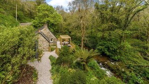 A stone cottage with a wooden deck and a small garden, surrounded by dense forest and a stream. - Romantic Riverside Cottage by the beach with Hot Tub, Sauna & Log Burner - Former Art Studio of Samu (Lamorna)