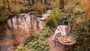 A rustic outdoor seating area with a table set for a meal, surrounded by a natural landscape and a small stream. - Romantic Riverside Cottage by the beach with Hot Tub, Sauna & Log Burner - Former Art Studio of Samu (Lamorna)