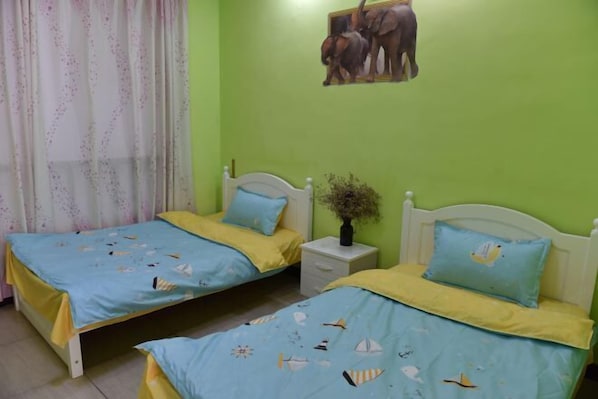 A room with two beds, each with a blue and yellow bedspread, a white nightstand with a vase, and a green wall with a picture of elephants.