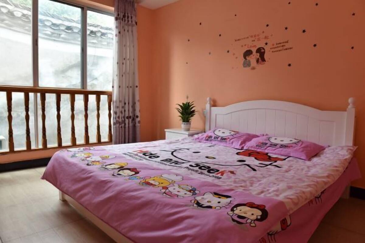 A bedroom with a pink bedspread featuring Hello Kitty characters, a white headboard, a nightstand with a plant, and a window with a view of a traditional building.