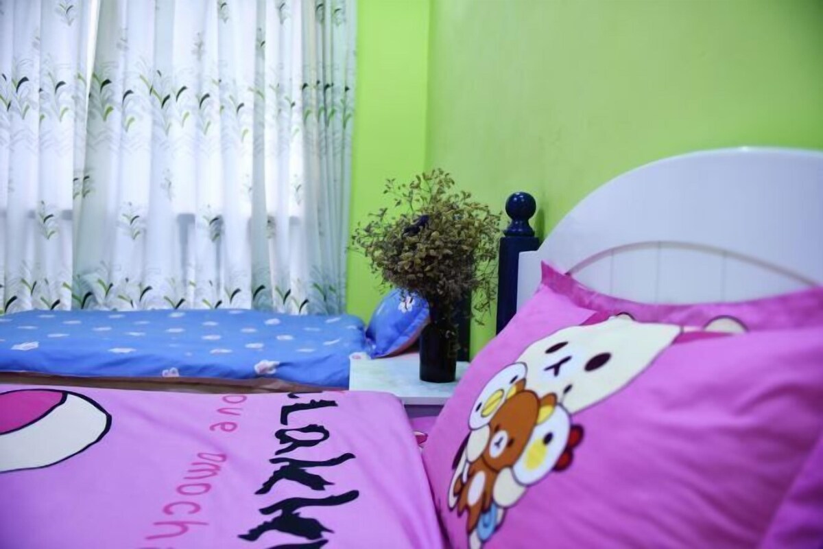 A bedroom with a bed, pillows, a nightstand, a vase with dried flowers, and a window with curtains.