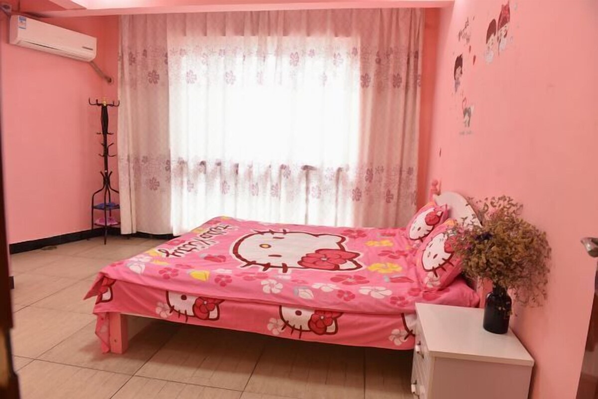A pink-themed bedroom with a Hello Kitty bedspread, a white bedside table, and a potted plant.