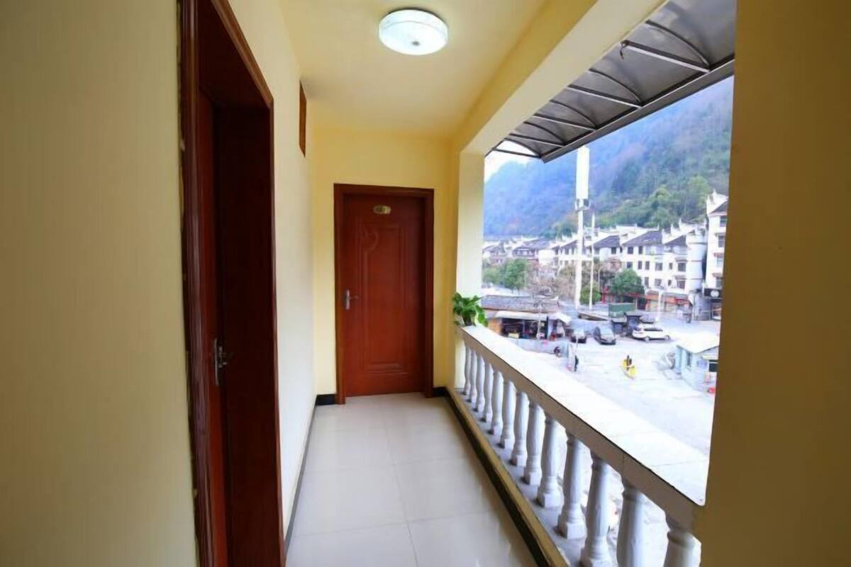 A corridor with a wooden door, a balcony with a railing, and a view of a town with buildings and mountains.