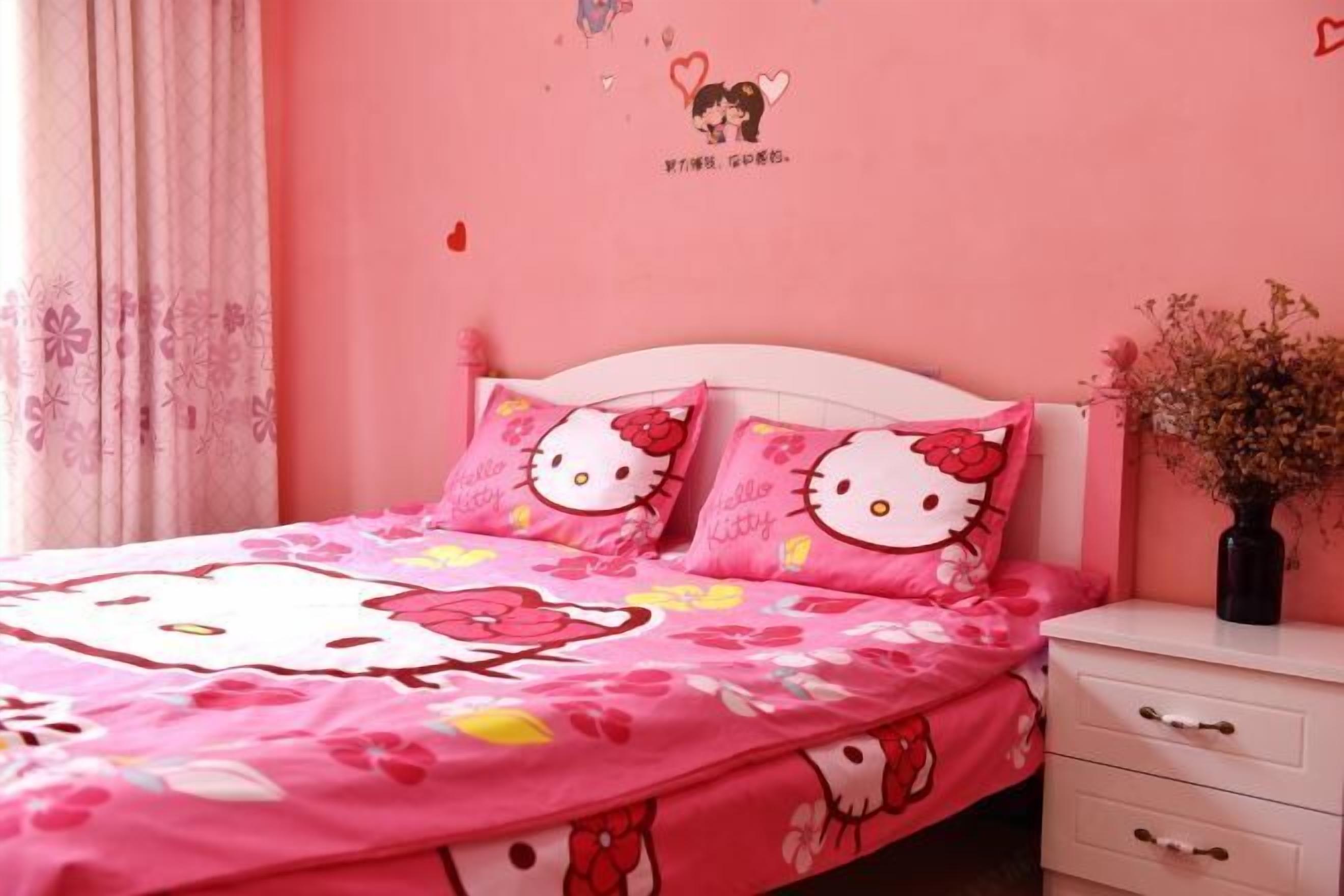 Un dormitorio con temática rosa, con una cama, almohadones de Hello Kitty, una mesita de noche blanca y un jarrón con flores secas.