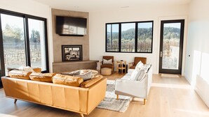 A spacious living room with a large sofa, a white armchair, and a fireplace. There are large windows offering a view of a vineyard and mountains. - Calistoga Vineyard House (Calistoga)