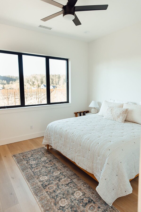 A bedroom with a large bed, a ceiling fan, a window with a view of a vineyard, and a patterned rug. - Calistoga Vineyard House (Calistoga)