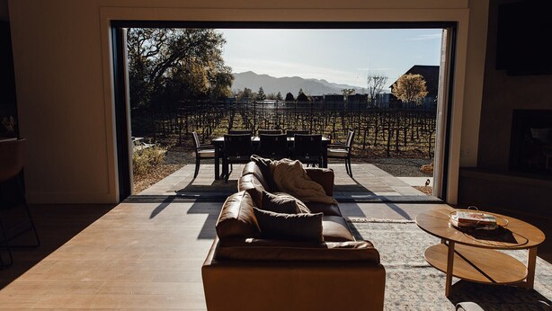 A modern living room with a large sliding glass door leading to an outdoor patio. Inside, there is a leather sofa, a coffee table, and a dining area with chairs. - Calistoga Vineyard House (Calistoga)