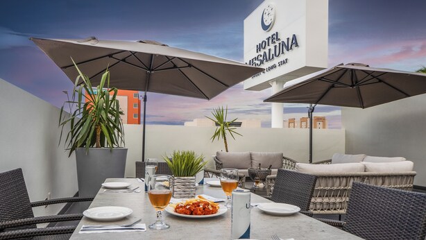 Un espace repas avec une table dressée pour deux personnes, des parasols, des meubles en osier et une vue sur l’enseigne de l’hôtel.
