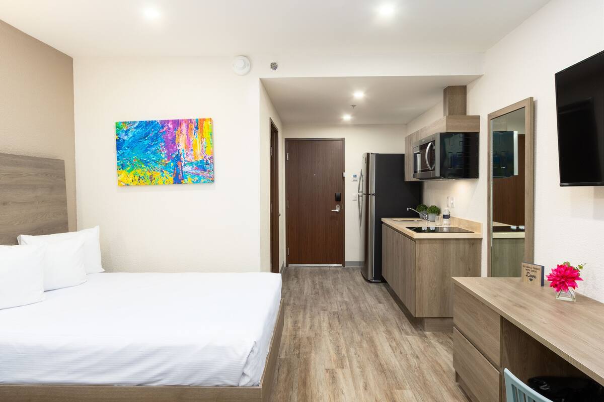 A hotel room with a bed, a colorful abstract painting, a kitchenette with a microwave, and a TV mounted on the wall.