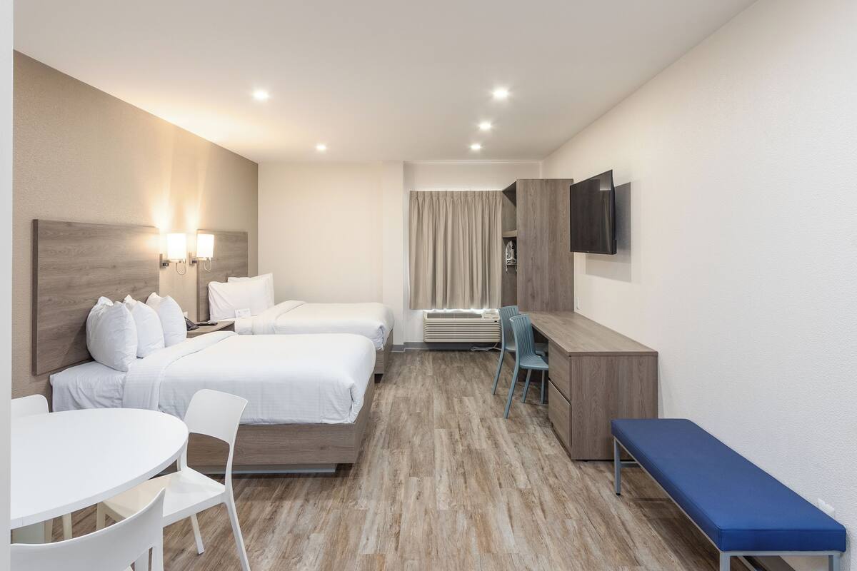 A hotel room with two beds, a desk, a chair, a bench, a TV, and a window with curtains.