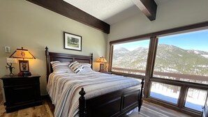 A bedroom with a large bed, wooden floor, and a view of snow-covered mountains through a large window. - Dreamcatcher - 2 bed 2 bath condo with tremdous views! (Estes Park)