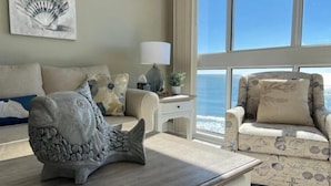 Ein Wohnzimmer mit großem Fenster zum Meer, eine Steinfischskulptur auf einem Tisch und ein gemustertes Sessel.