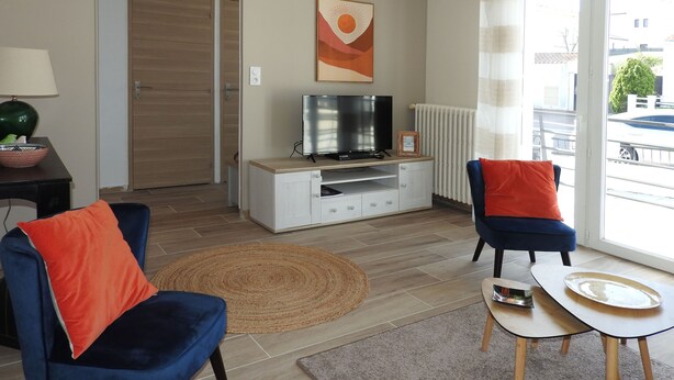 Un soggiorno moderno con una TV su un mobile in legno, due poltrone con cuscini arancioni, un tavolino con un vassoio e un'ampia finestra vista esterna.