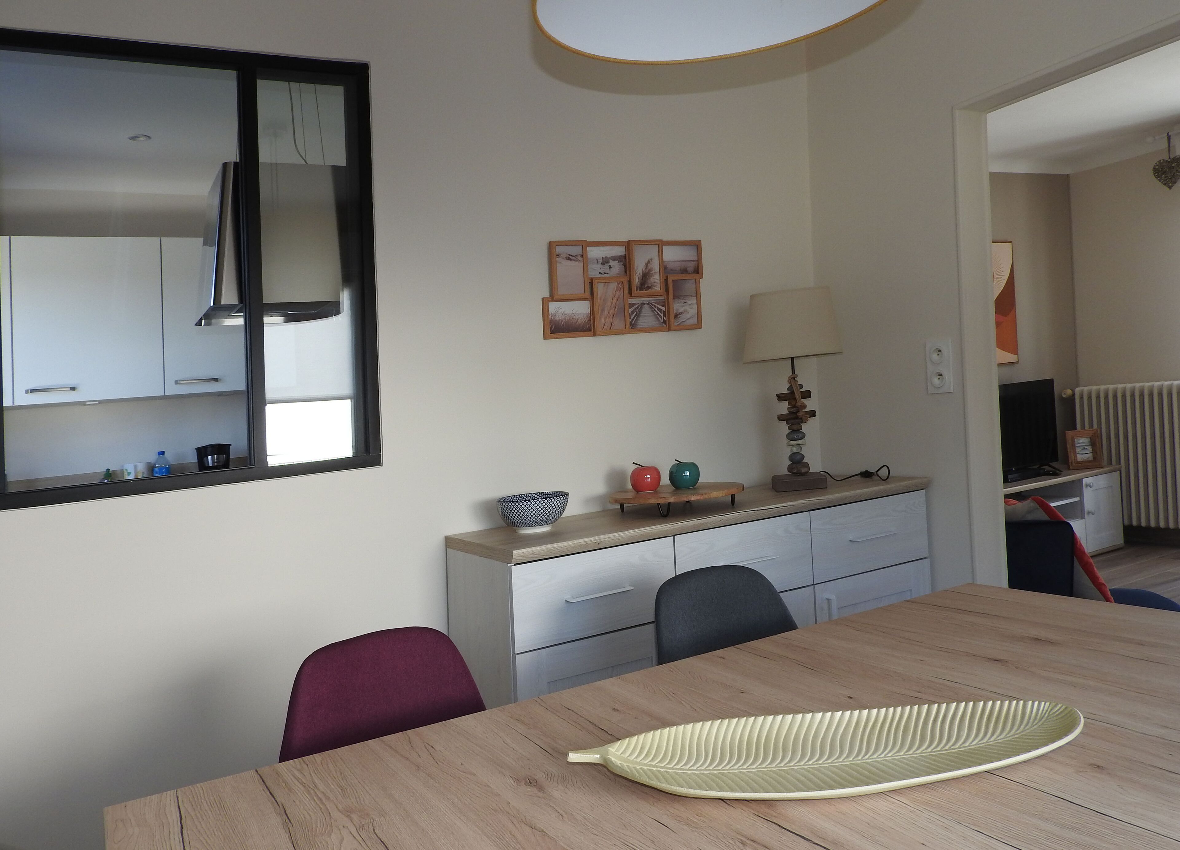 Uma sala de jantar com mesa de madeira, cadeiras, um espelho, um pequeno armário com objetos decorativos e uma televisão ao fundo.
