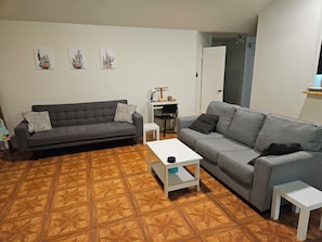A living room with two sofas, a coffee table, and a small white table. There are three wall-mounted artworks and a door leading to another room.