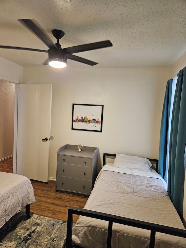 A bedroom with two beds, a ceiling fan, a dresser, and a cityscape picture on the wall.