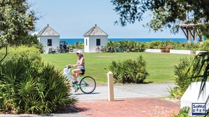 En kvinde cykler på en asfalteret sti med udsigt til havet og to små bygninger i baggrunden.