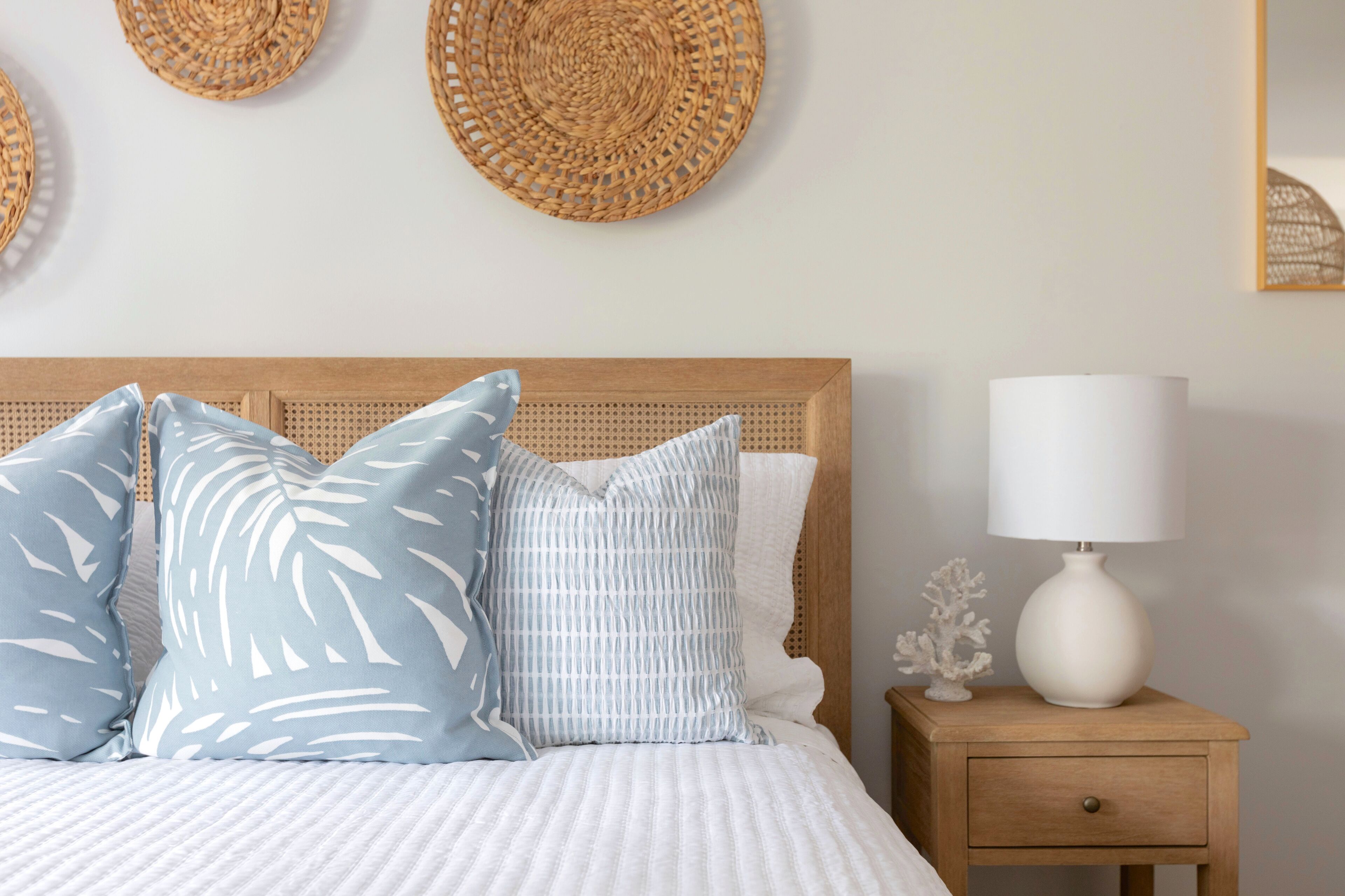 A neatly made bed with a white comforter and blue patterned pillows, a wooden nightstand with a white lamp, and woven wall decorations.